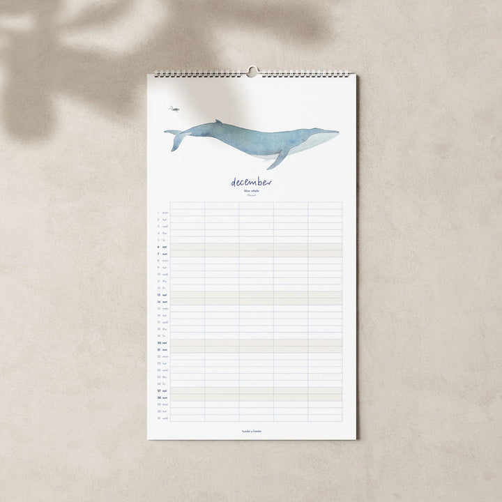 December page of the 2025 family planner calendar featuring a blue whale illustration above a grid layout for organizing family events and monthly schedules.