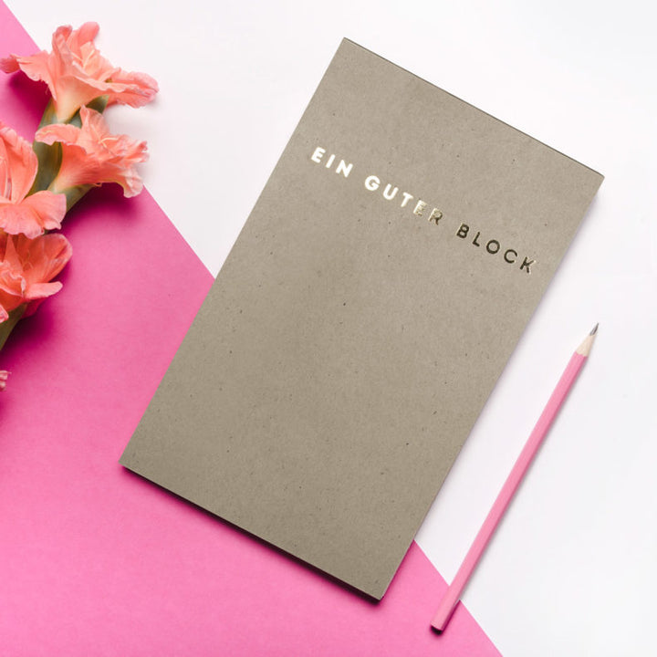 Stylized image of "Ein Guter Block" notepad placed on a pink background with flowers and a pink pencil.